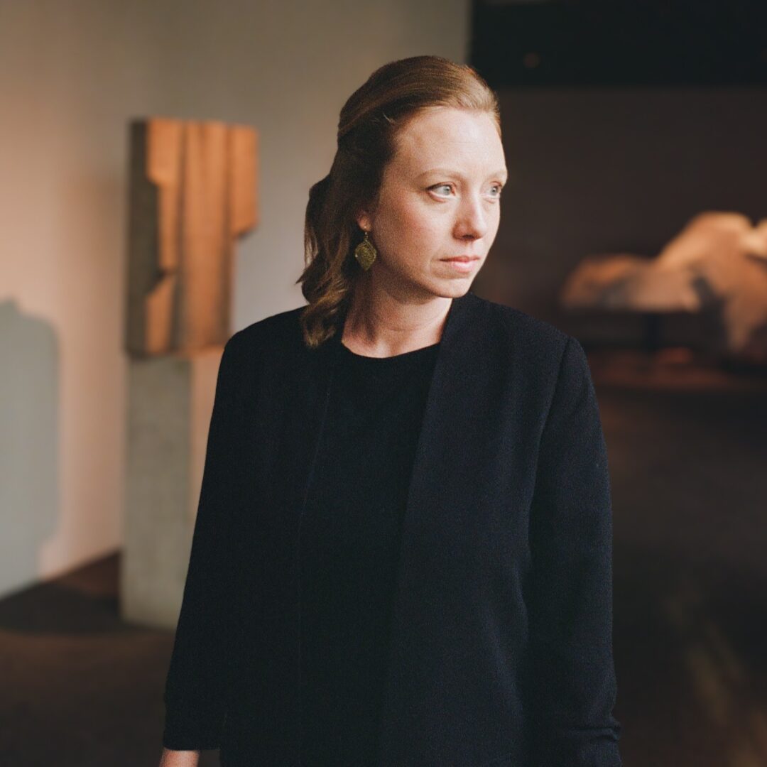 A woman in a black outfit looks thoughtfully to the side in a softly lit room.