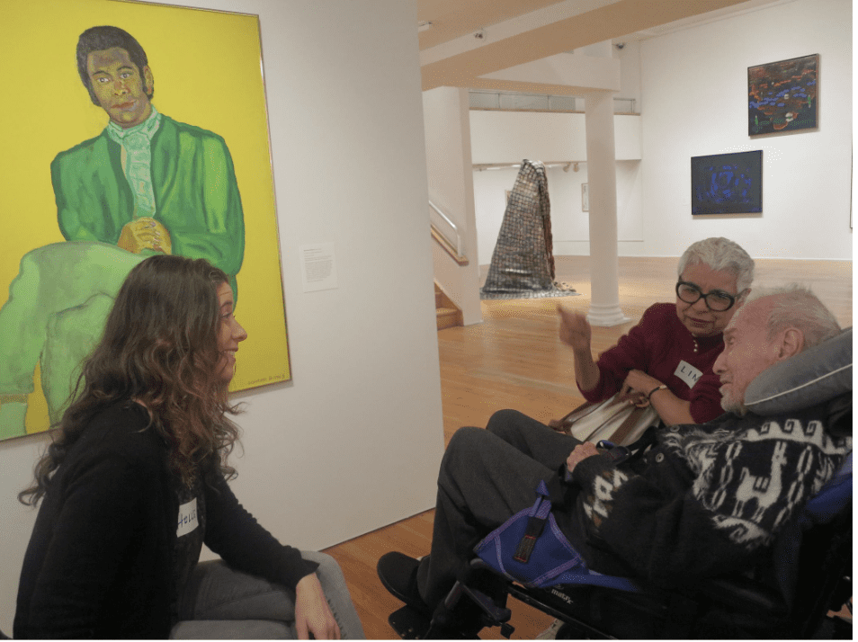 Two women conversing in an art gallery with paintings in the background.